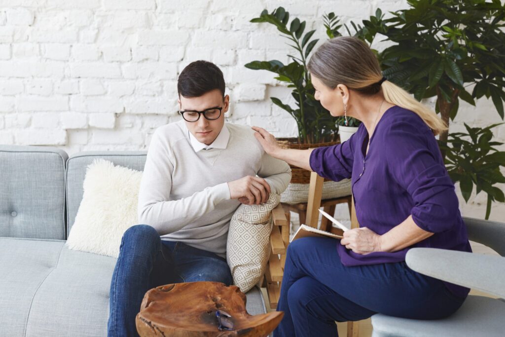 Candid shot of casually dressed professional woman psychotherapist in her fifties touching her young male patient by shoulder while having counseling session